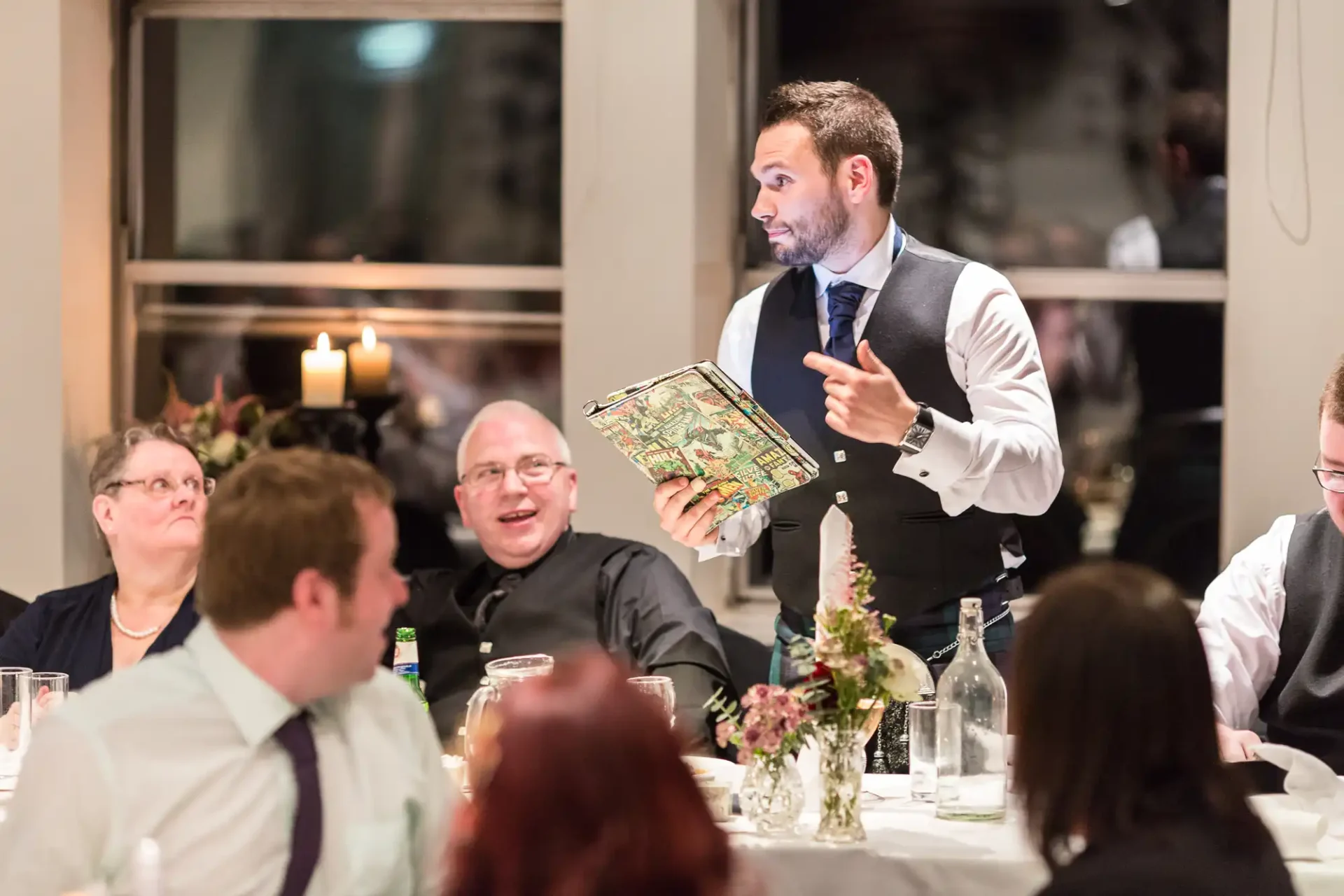 A waiter presenting a menu to diners at an elegantly set table in a restaurant, engaging in discussion with a seated couple.
