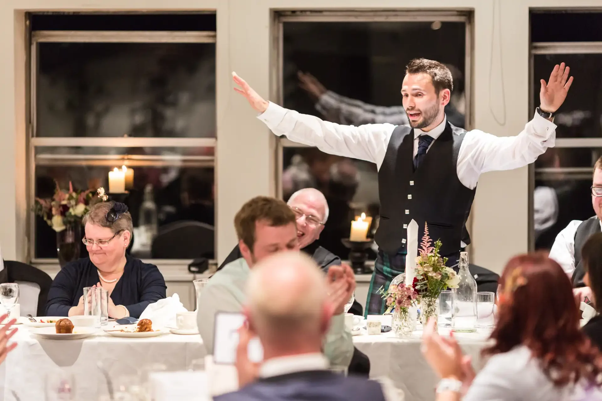 A man in a vest and tie gestures with arms wide open, speaking to seated guests at a warmly lit dinner event.