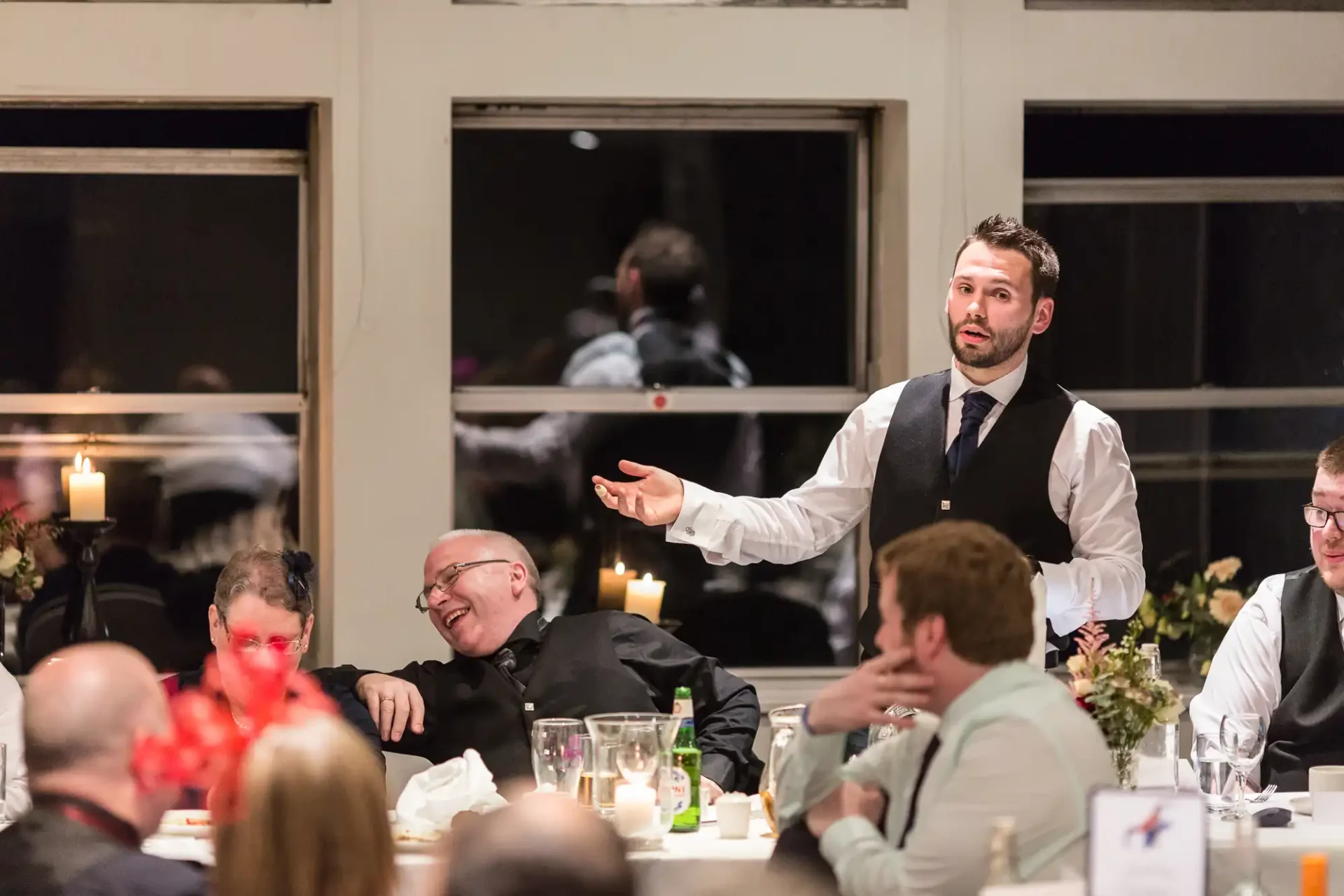A man in a vest and tie gesturing while speaking at a dinner event, with guests seated at tables, some laughing and engaging with him.