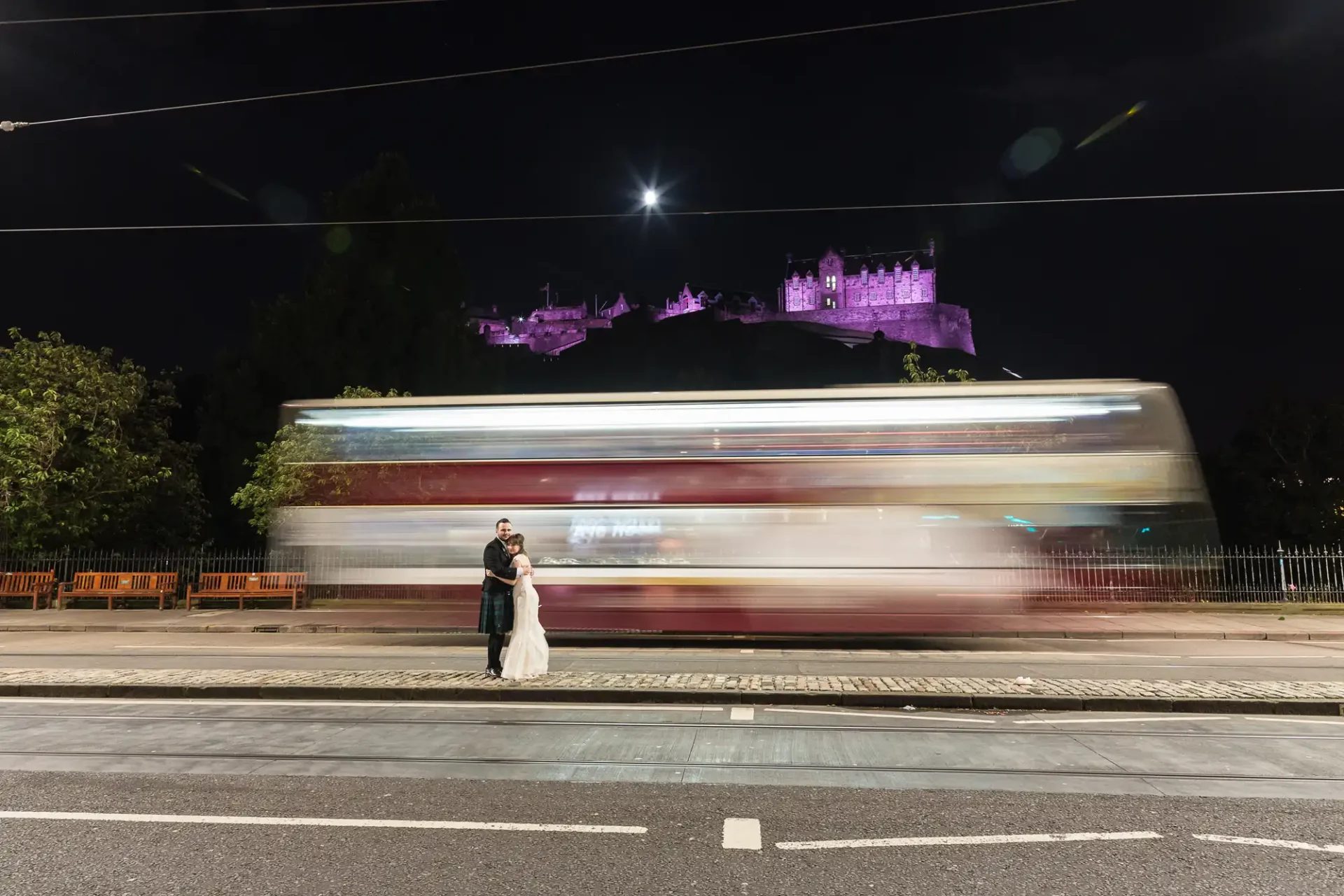 A couple embracing on a city street at night, with a blurred tram passing by and a lit-up castle on a hill in the background.