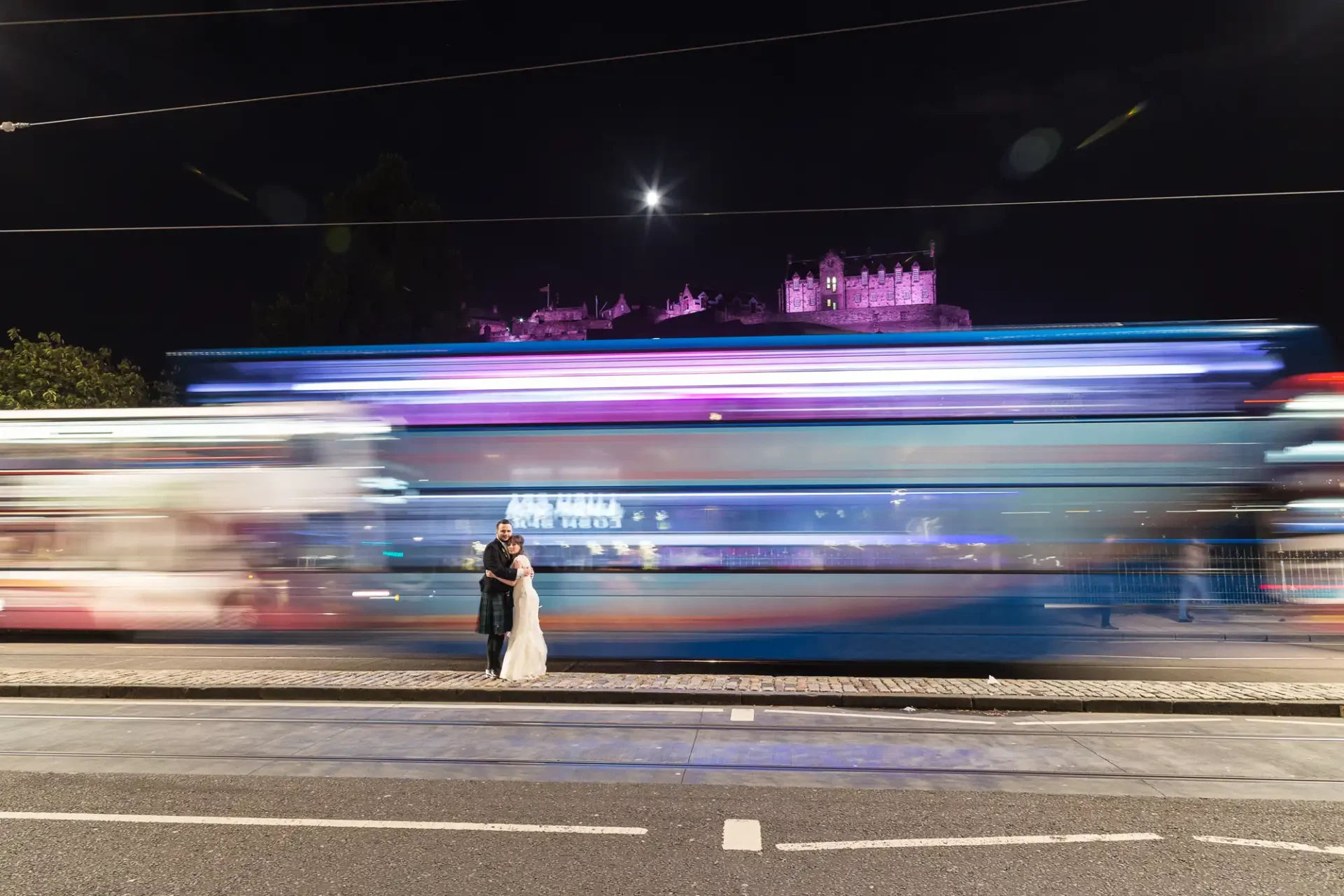 A couple embraces on a city street at night with a motion-blurred bus passing by and a lit castle in the background.