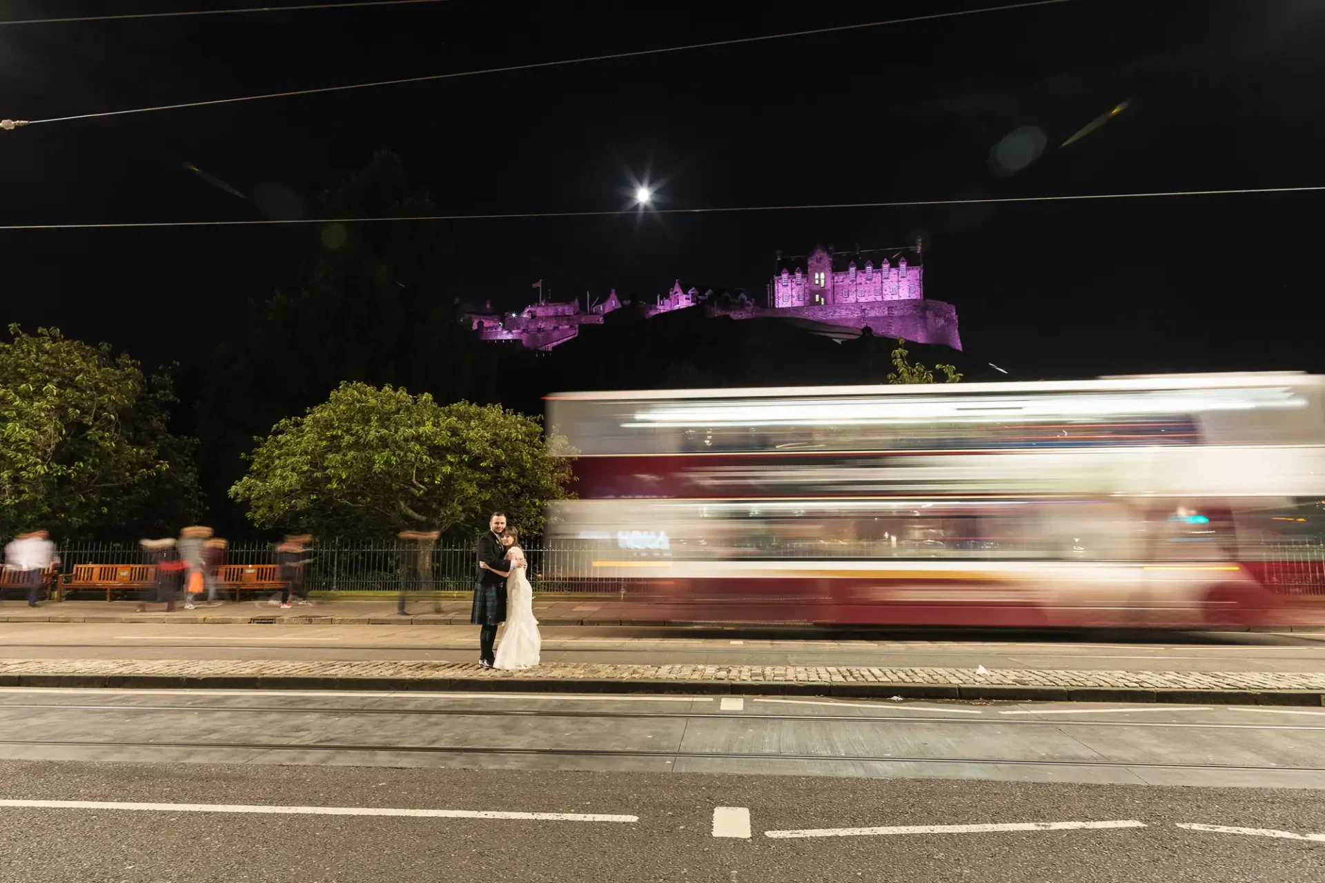 Night view of a couple holding hands on a street with a blurred bus passing by, and edinburgh castle illuminated in the background.