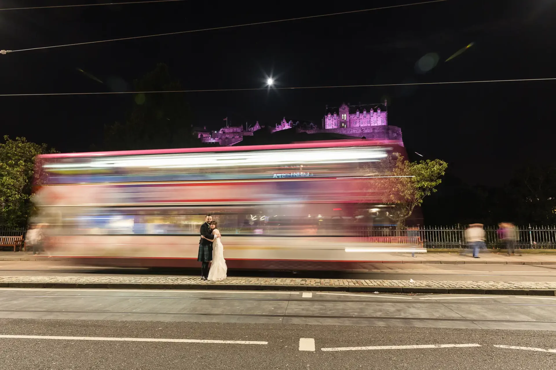 A couple embracing in front of a blurred city bus at night with an illuminated castle on a hill in the background.