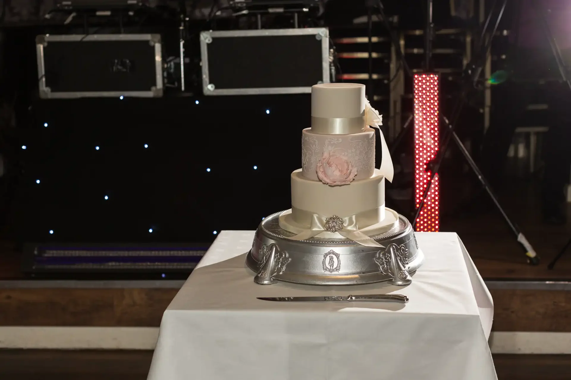 A three-tiered wedding cake on a silver stand, set against a dark backdrop with sparkling lights and stage equipment.