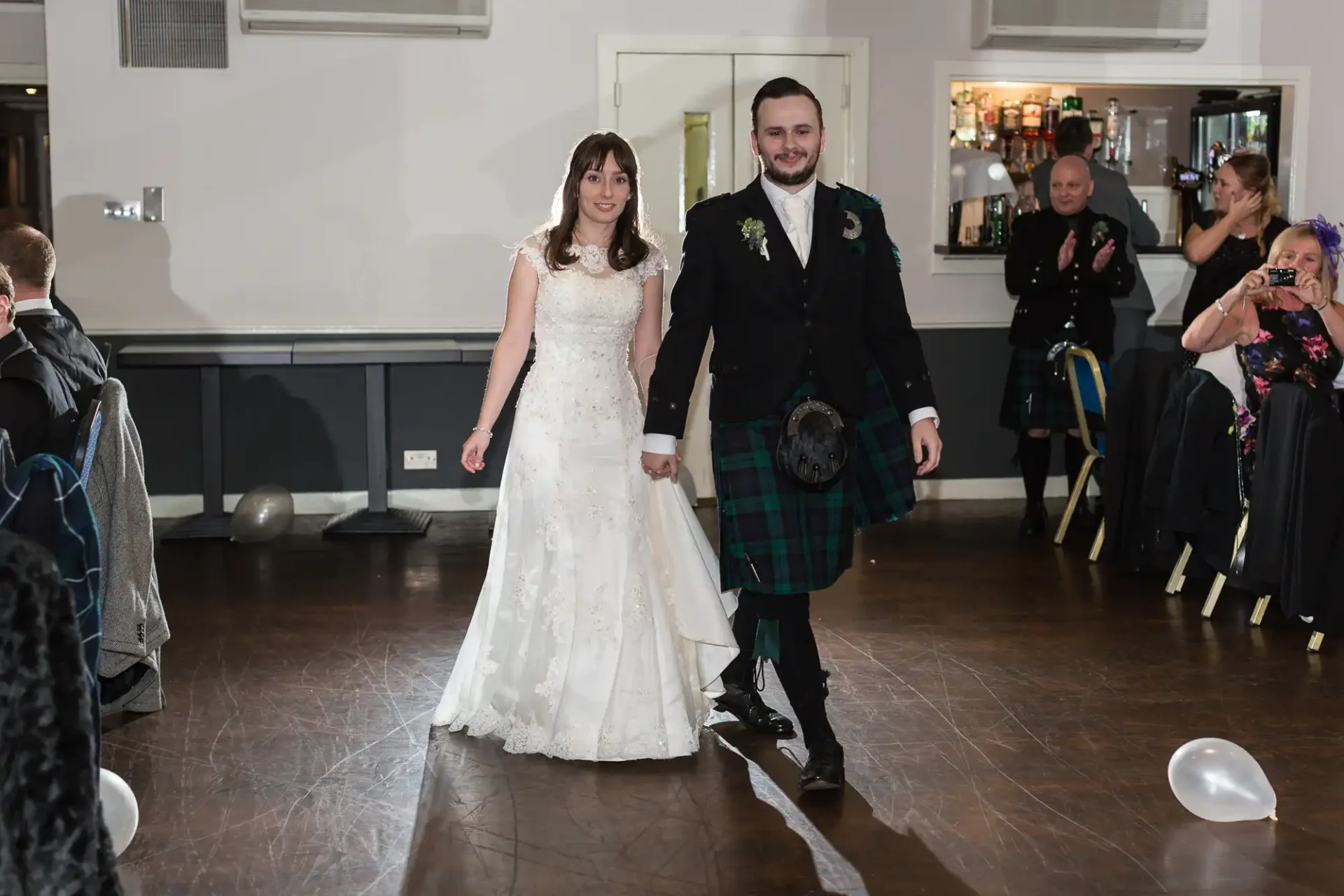 A bride in a white gown and a groom in a kilt walk hand-in-hand at a wedding reception, with guests looking on.