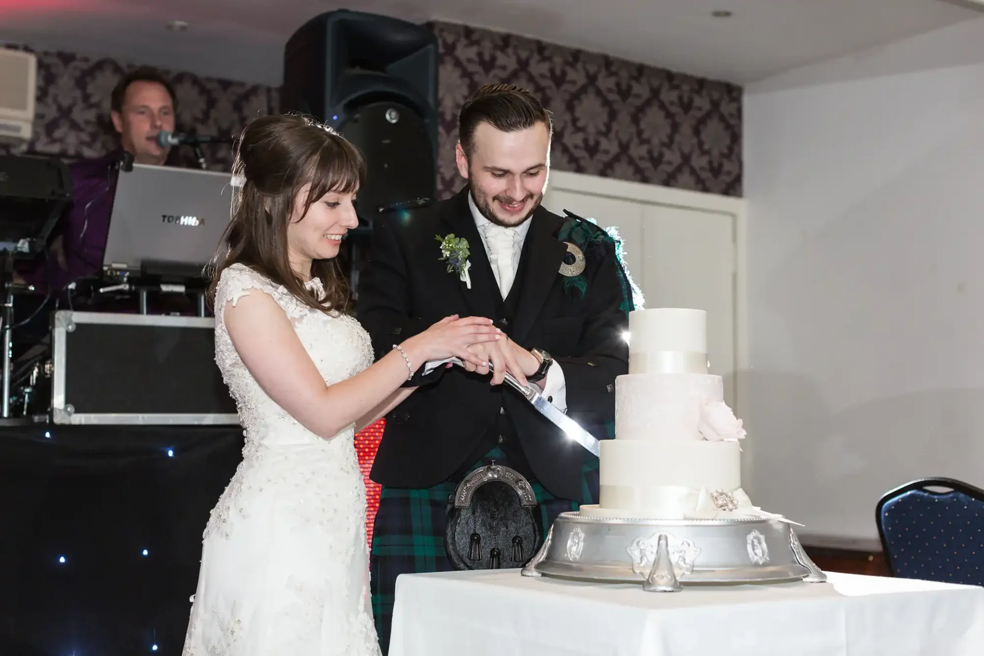 A bride and groom cutting their wedding cake together, smiling, with a dj in the background.