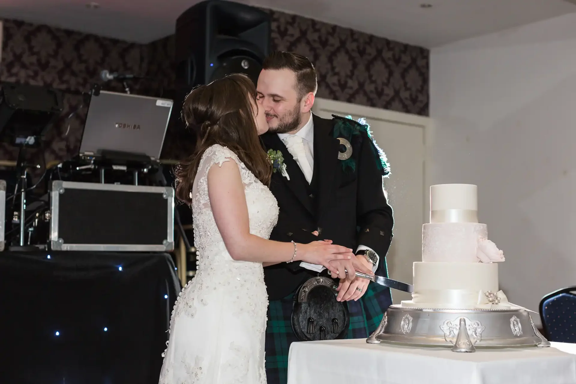 A bride and groom in a kilt cutting a wedding cake together at a reception.