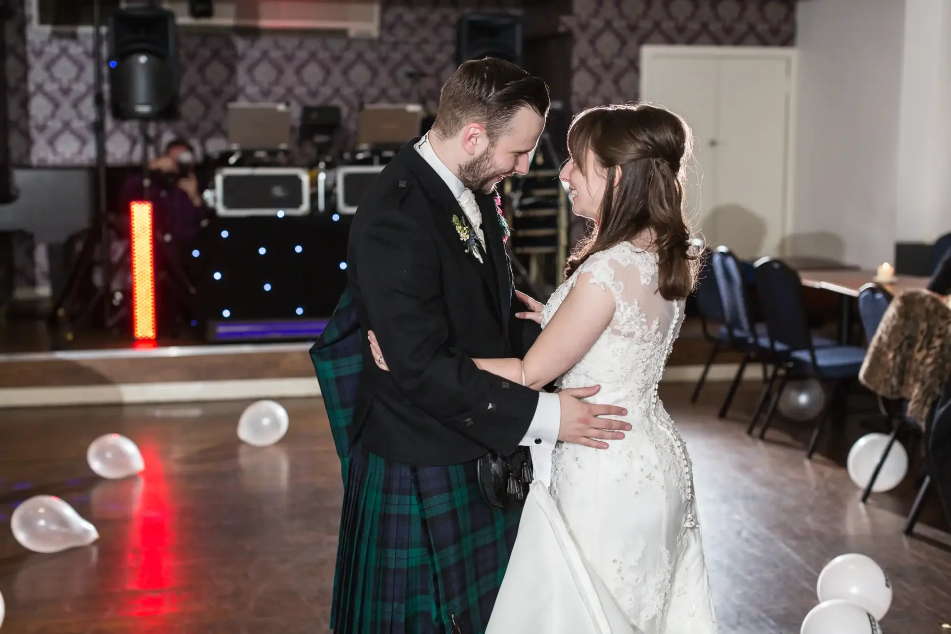 A bride and groom share a romantic dance, the groom in a tartan kilt and the bride in a white dress, in a ballroom with dim lighting and scattered white balloons.