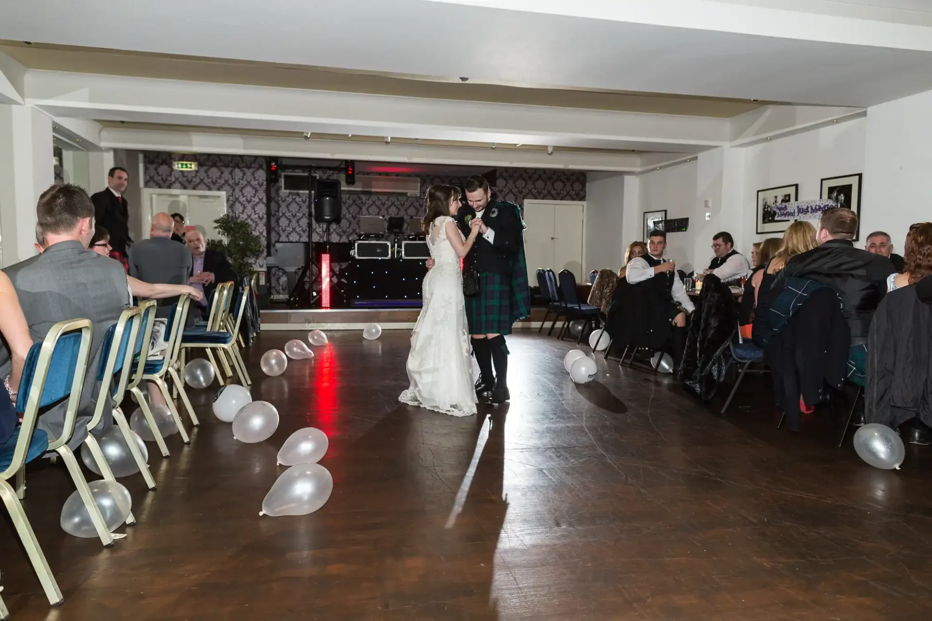 A bride and groom share their first dance in a decorated hall with guests watching and white balloons on the floor.