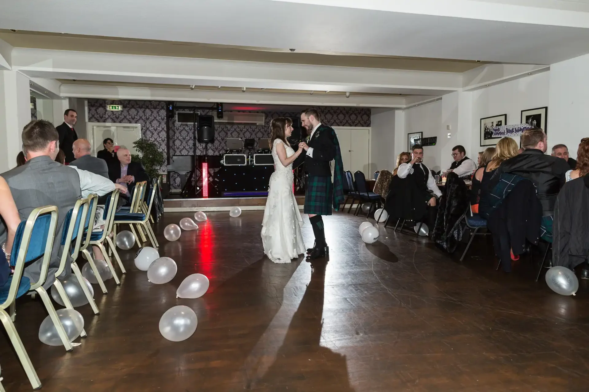 A bride and groom share their first dance in a hall decorated with balloons, surrounded by seated guests watching them.