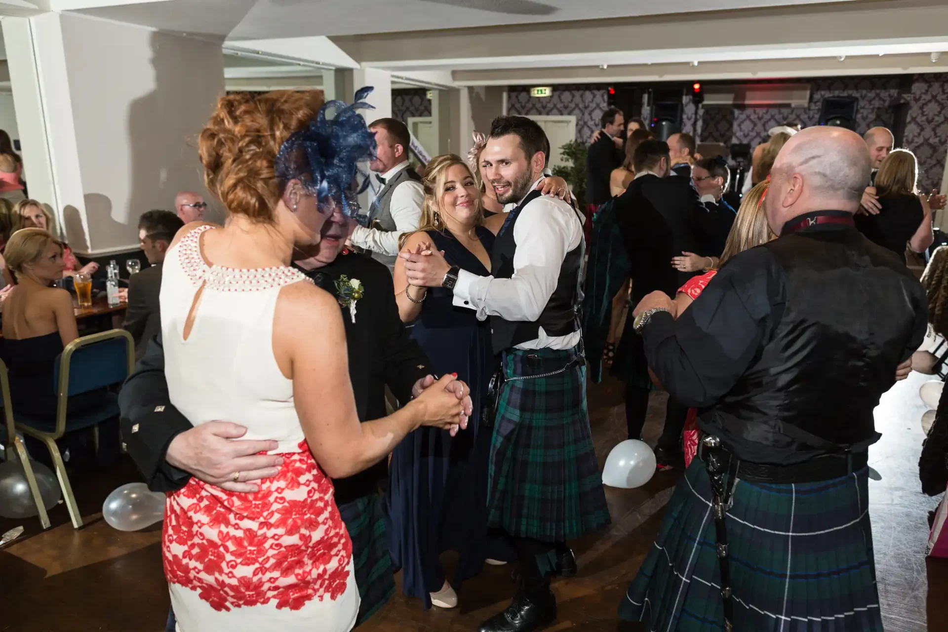 People in formal attire, including kilts, dancing at an indoor celebration with balloons and festive decor.