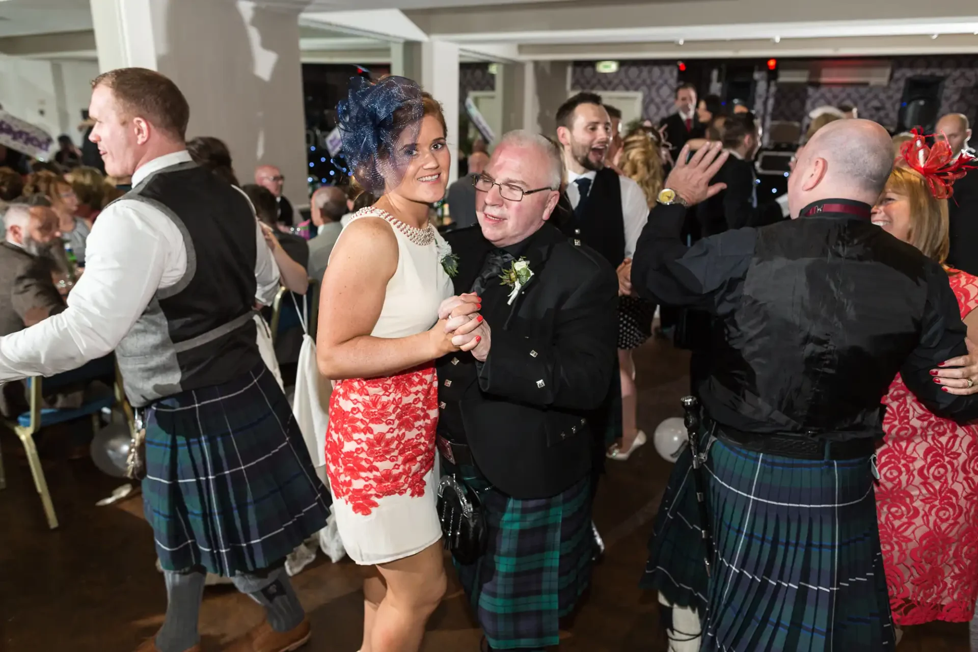 A couple in semi-formal attire, including kilts, dances joyfully among other guests at an indoor celebration.