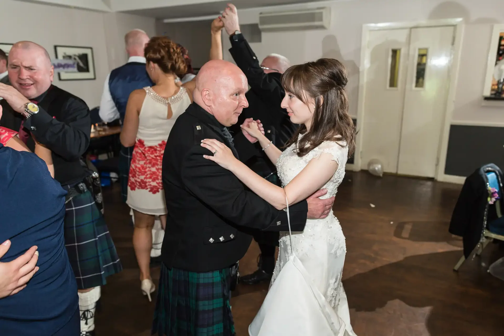 A bride in a white dress dancing closely with a bald man in a dark jacket and kilt at a lively indoor wedding reception.