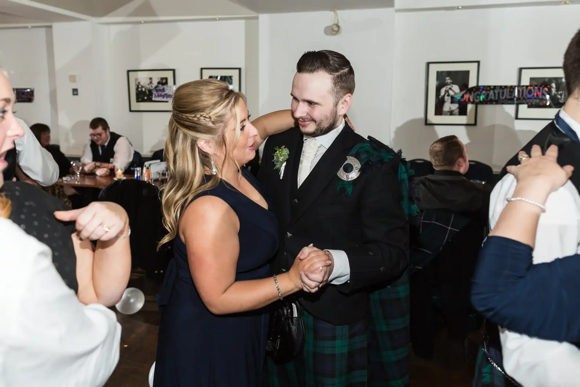 A man in a kilt and a woman with a corsage dancing and smiling at each other at a lively indoor celebration.