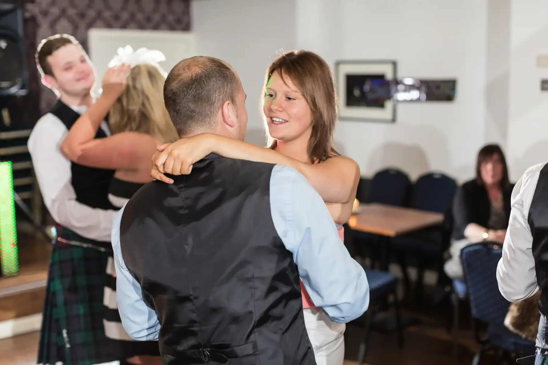 A couple dancing closely at an indoor event, with other guests and a man in a kilt visible in the background.