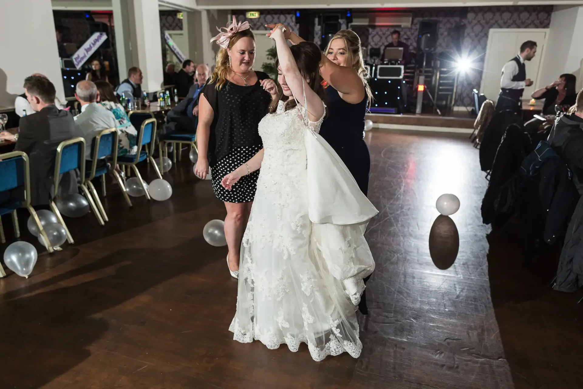 Three women dancing joyously at a wedding reception, two assisting the bride with her veil on the dance floor.