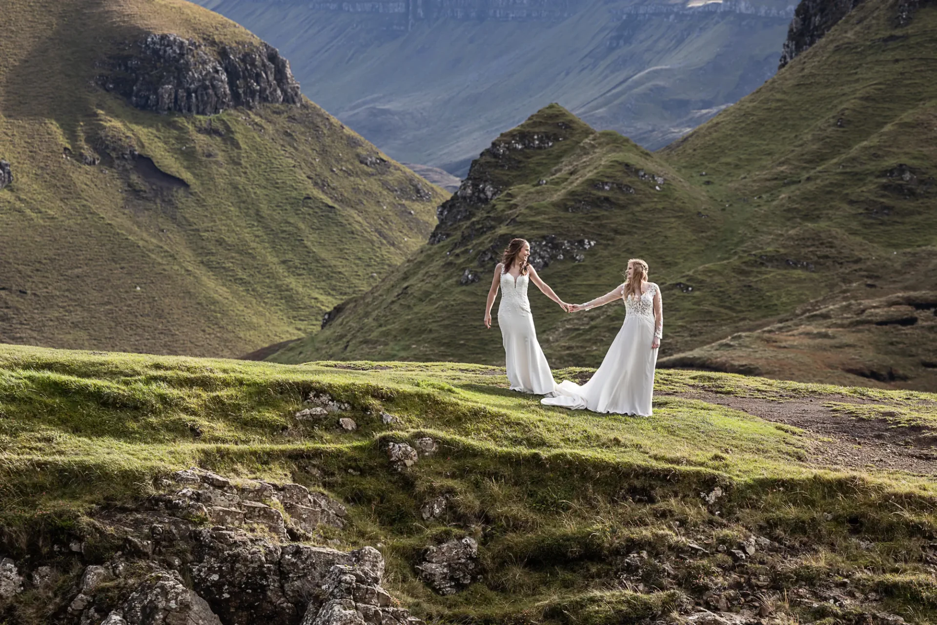 Two women in white dresses hold hands while walking on a grassy, mountainous terrain under a cloudy sky.