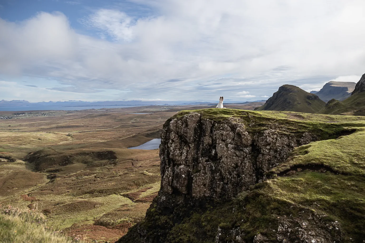 newlyweds overlooking the Isle from The Quiraing, standing on a grassy cliff overlooking a vast landscape with distant mountains under a partly cloudy sky at The Quiraing, Isle of Skye.