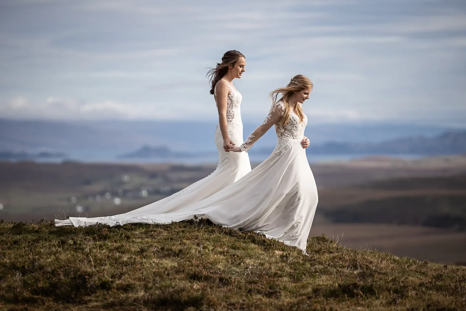 Scottish elopements: Two brides in white wedding dresses hold hands while walking on a grassy hill with a scenic landscape in the background at The Quiraing, Isle of Skye.