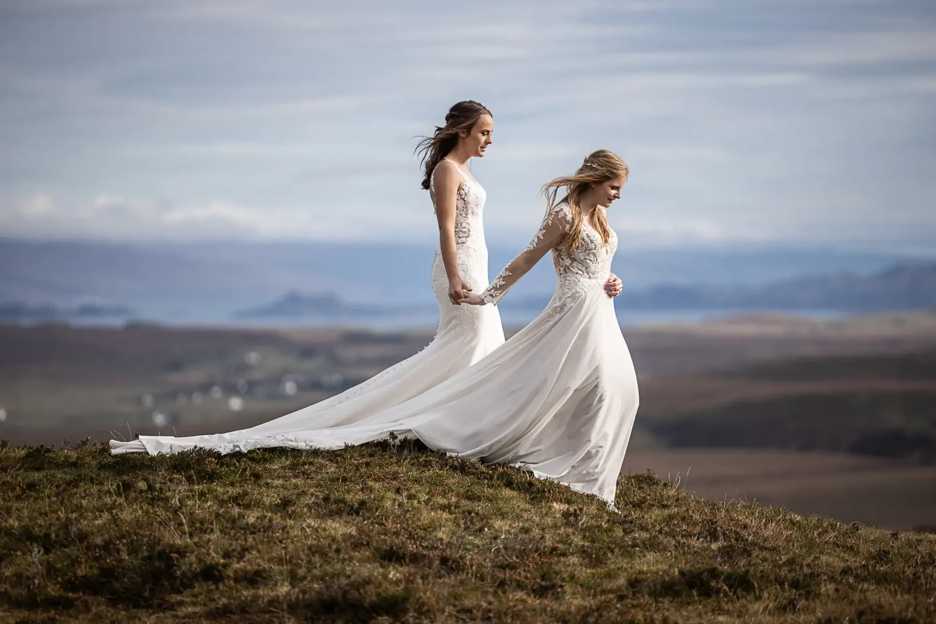 Two brides in white wedding dresses hold hands while walking on a grassy hill with a scenic landscape in the background at the Quiraing on Skye.