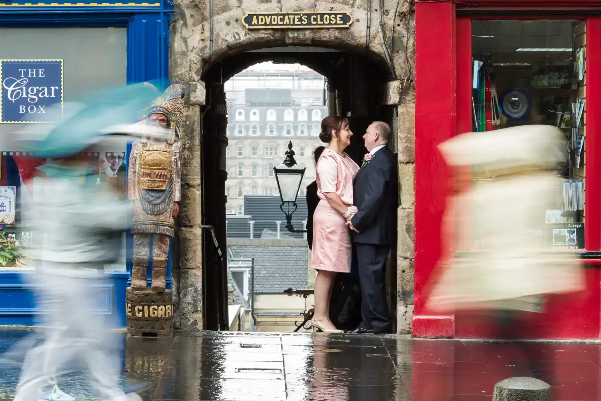 A couple stands facing each other under an archway labeled "Advocate's Close" as blurred pedestrians walk past on a rainy street.