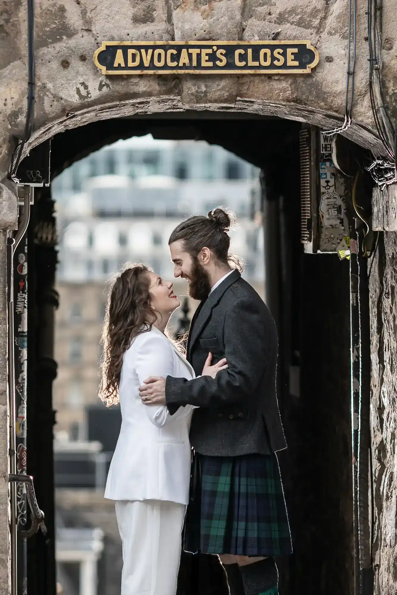 A couple stands embracing under an archway labeled "Advocate's Close"; the man wears a kilt and the woman is dressed in a white suit.