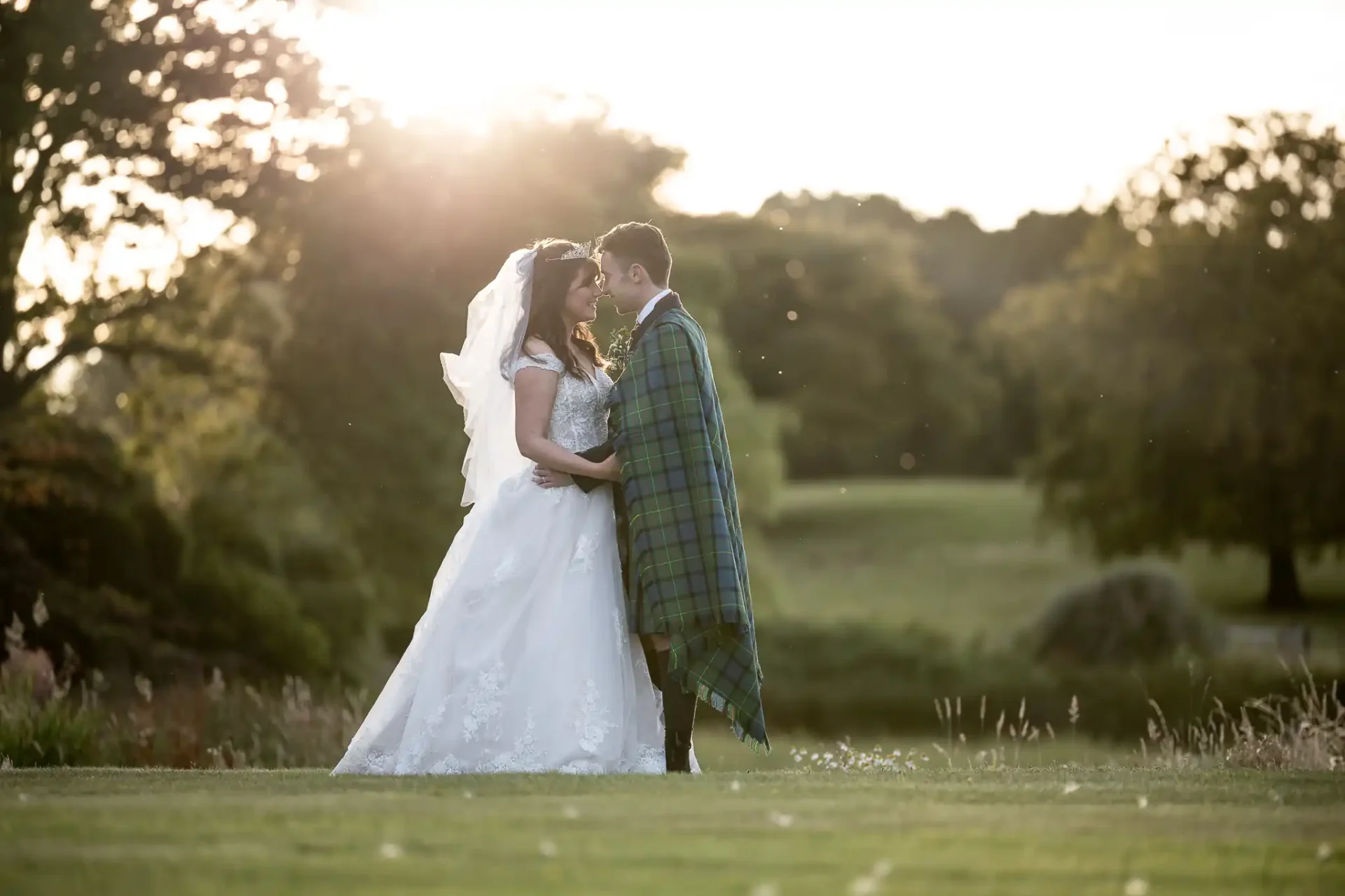 Another bride and groom who loved our wedding photography prices stand in a grassy field at sunset, with the groom wearing a plaid cloak.