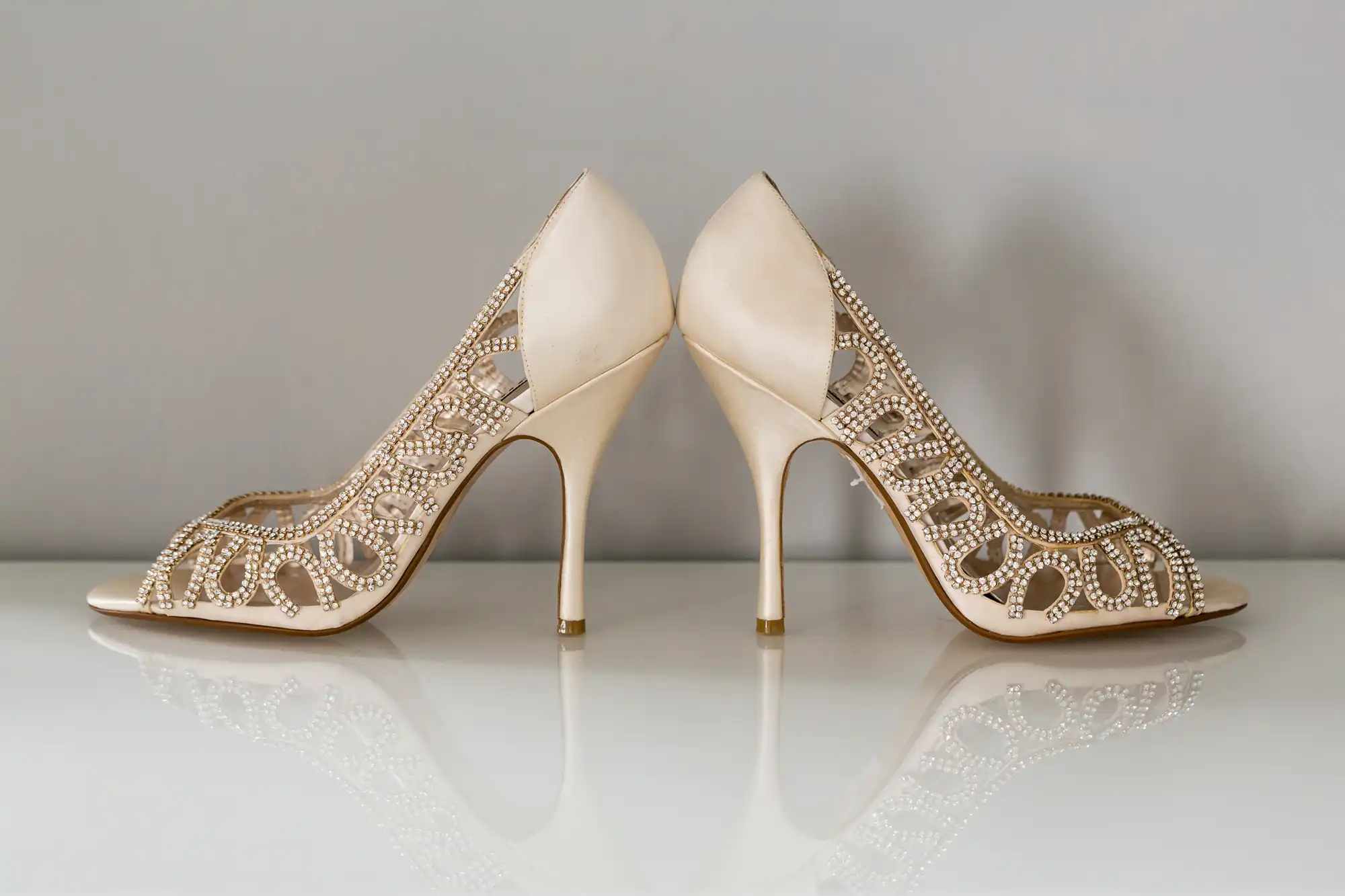 A pair of elegant beige high-heeled shoes with intricate golden embellishments, displayed on a reflective surface.