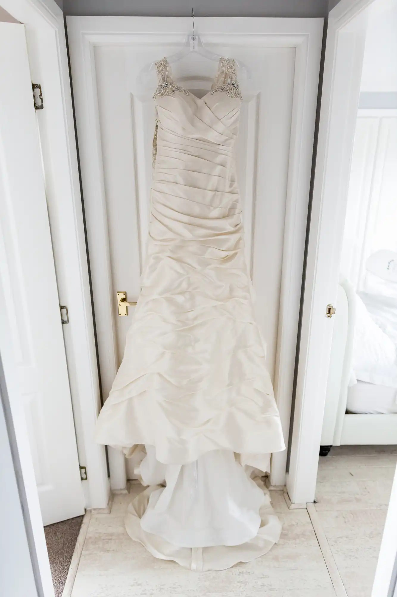 A white bridal gown with lace shoulder straps and ruched detailing, hanging on a white door in a bright room.