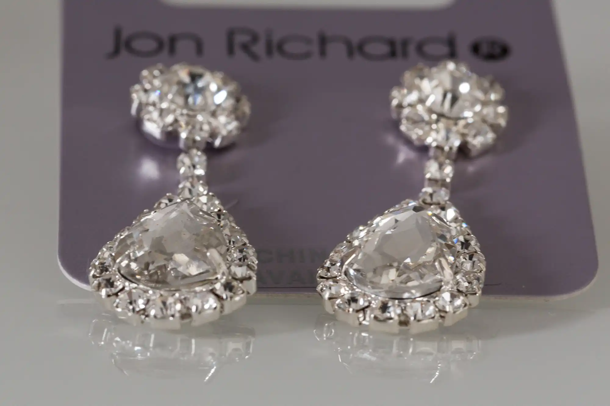 A pair of jon richard teardrop earrings with clear gemstones and silver settings, displayed on a branded card.