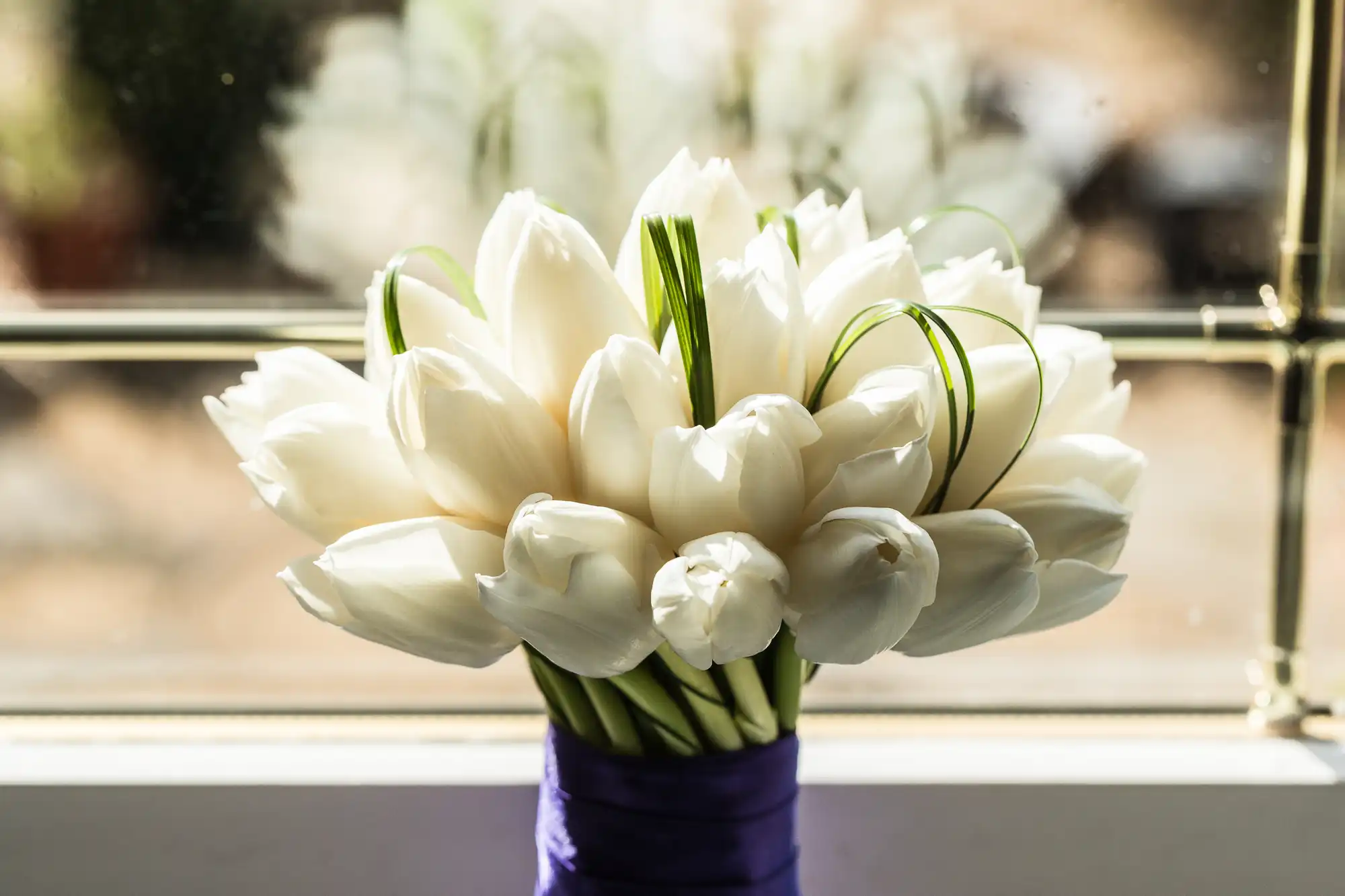 A bouquet of white tulips bound with a purple ribbon, positioned on a windowsill against a blurry background.