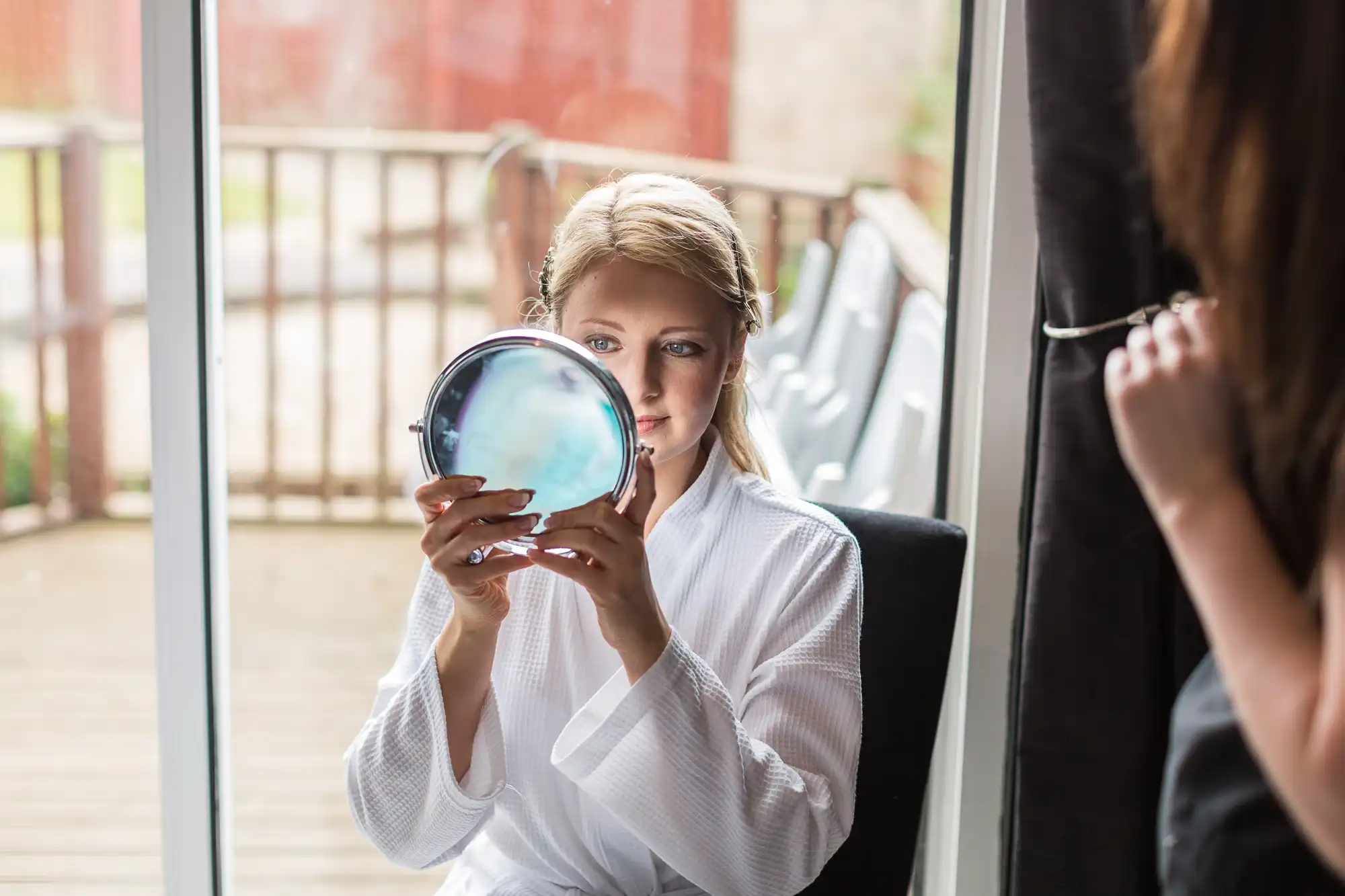 A woman in a white robe holding a mirror and applying makeup, sitting near glass doors with a reflection visible.