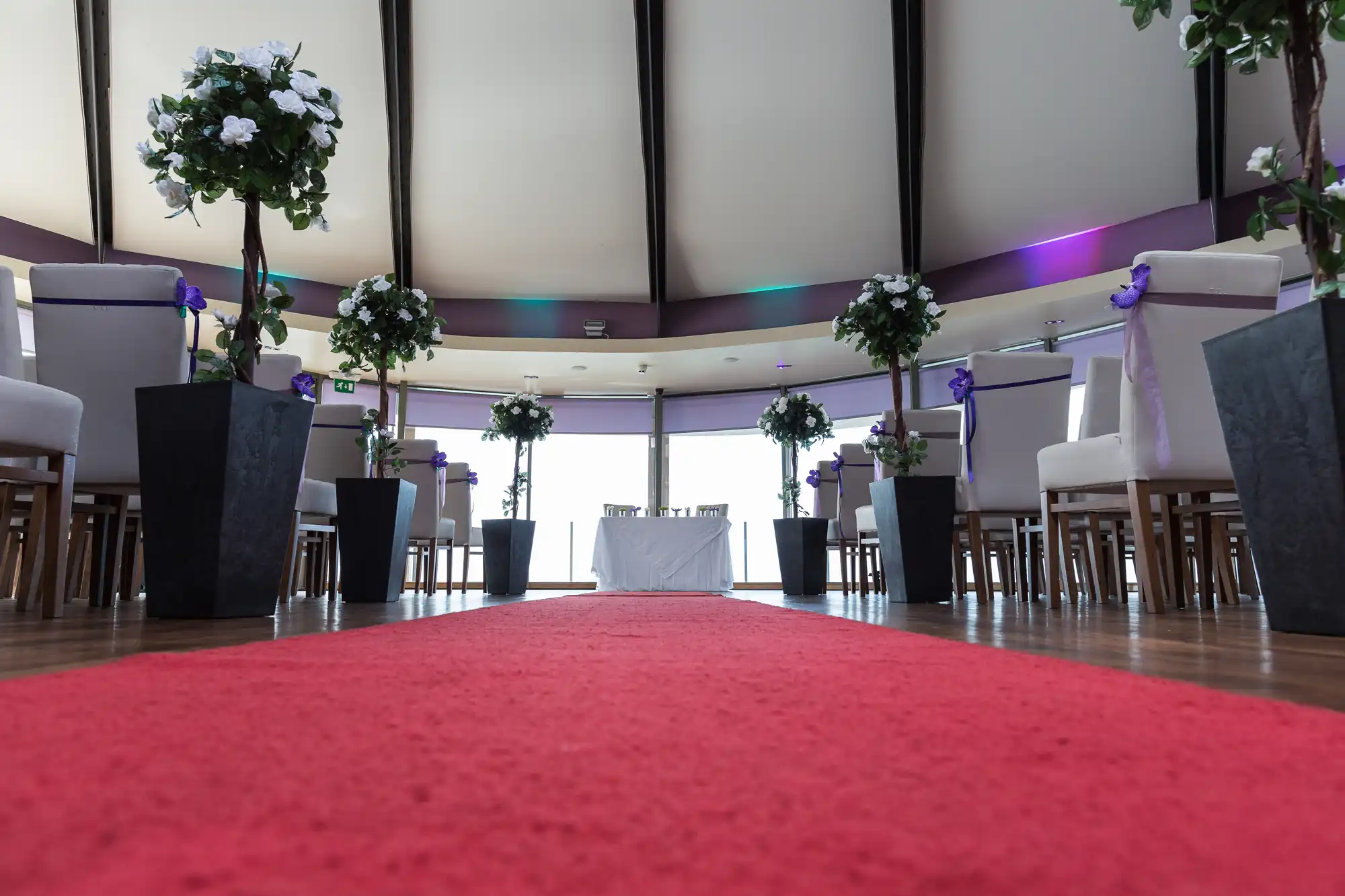 Interior of a wedding venue with red carpet aisle, white chairs adorned with purple ribbons, and floral decorations under soft lighting.