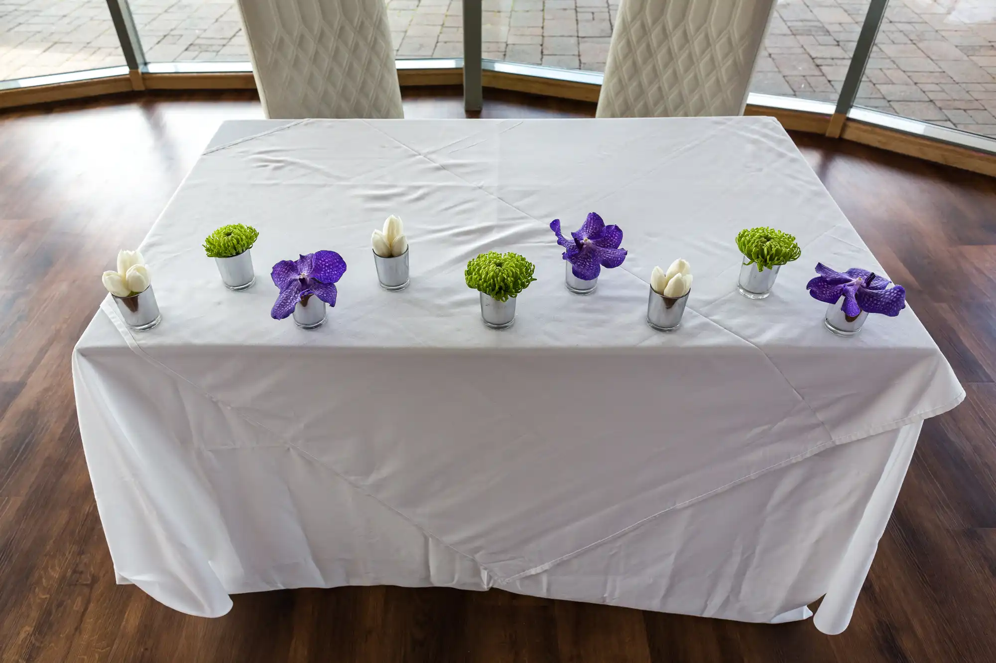 A rectangular table covered with a white tablecloth, decorated with symmetrically placed purple orchids and green plants in white pots, in a room with large glass windows.