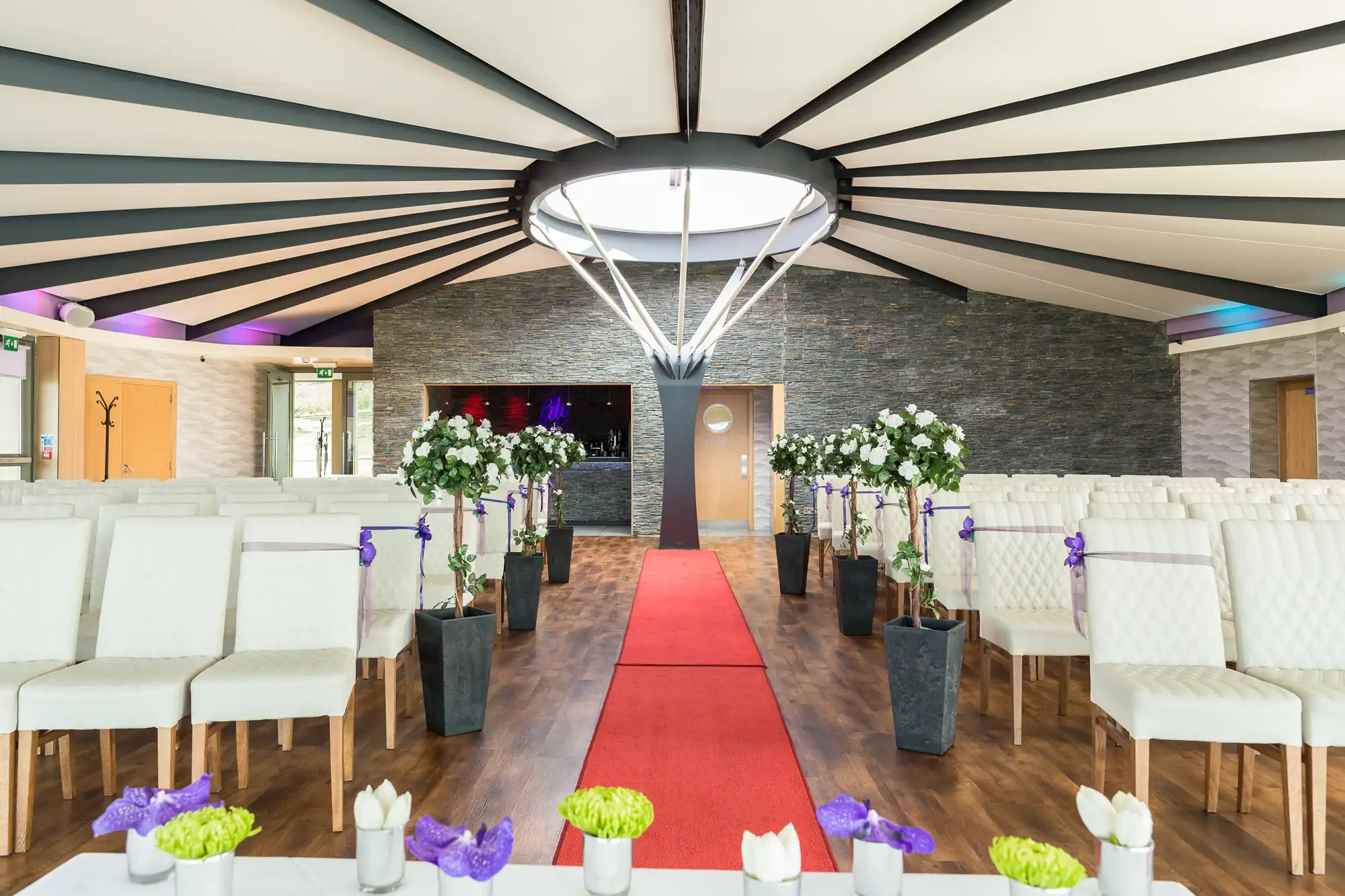 Interior of a wedding venue with white chairs, a red carpet aisle, and floral decorations under a modern ceiling with hanging chandelier.