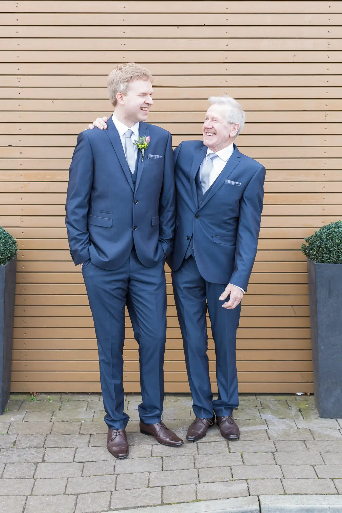 Two men in navy suits smiling at each other, standing against a wooden slat wall, with one man wearing a boutonniere.