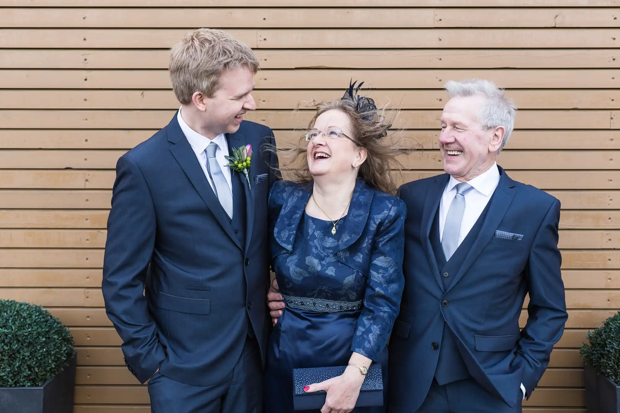 Three people laughing joyously at an event; a younger man in a suit on the left, a middle-aged woman in a blue dress and fascinator in the middle, and an older man in a suit on the right.