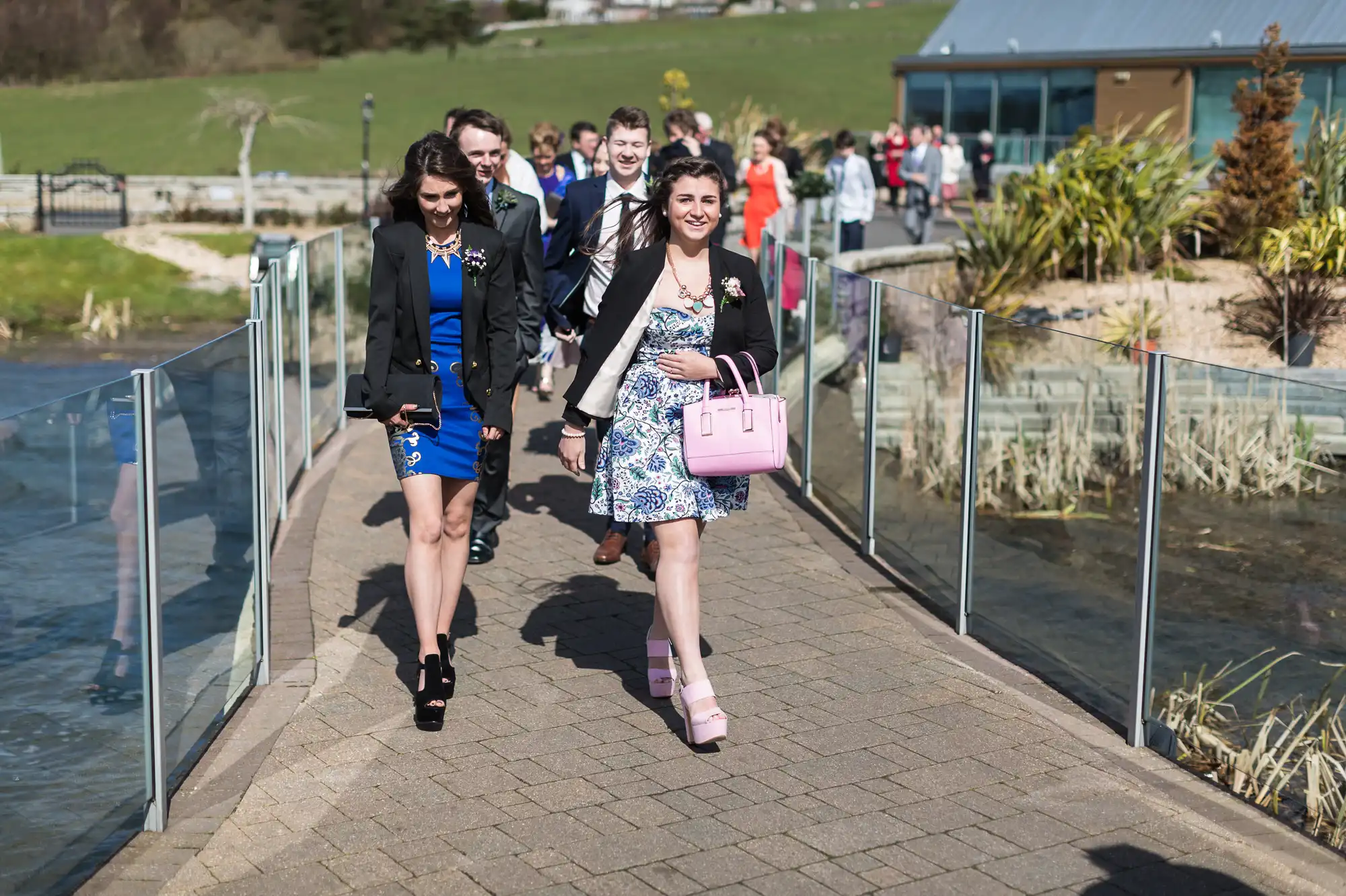 Group of people dressed in formal attire walking along a pathway at an outdoor event, with smiles and enjoying sunny weather.