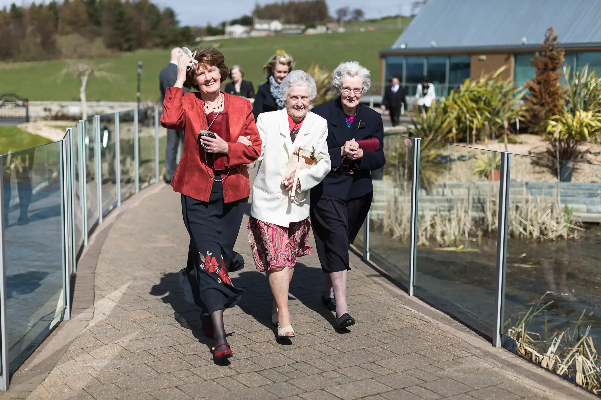 Three elderly women walking joyfully along a pathway beside a pond on a sunny day, one using a walking stick.