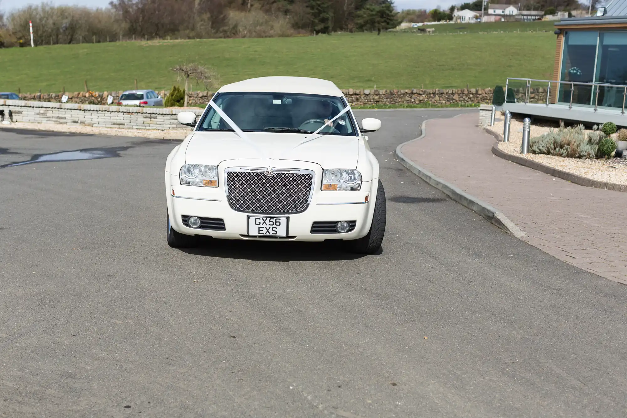 A white chrysler 300 parked on a tarmac driveway, with green hills and a building in the background.