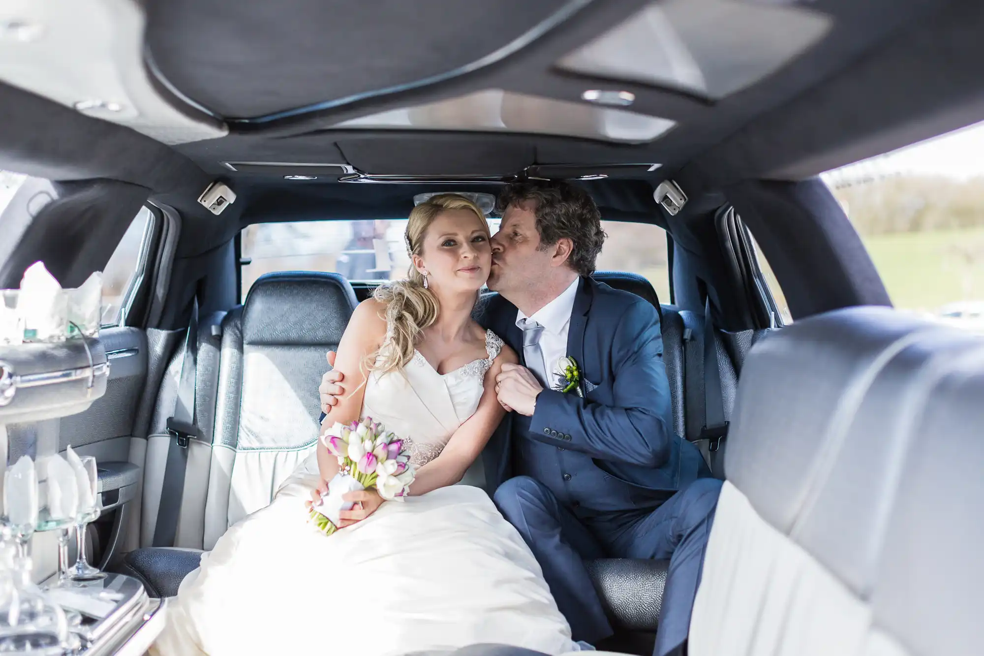 A groom in a blue suit kisses a bride in a white dress inside a spacious car, both holding bouquets.