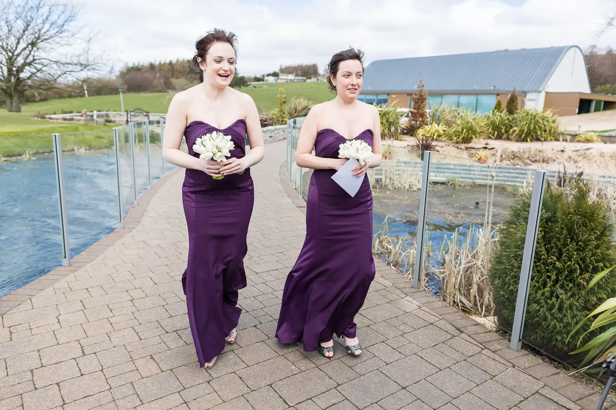 Two women in purple dresses holding bouquets walk along a paved path near a water feature, with a building in the background.