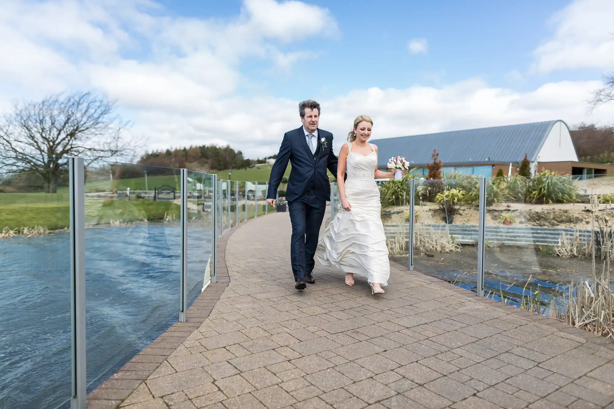 A bride and groom walking along a glass barrier-enclosed pathway with a pond and rural buildings in the background.