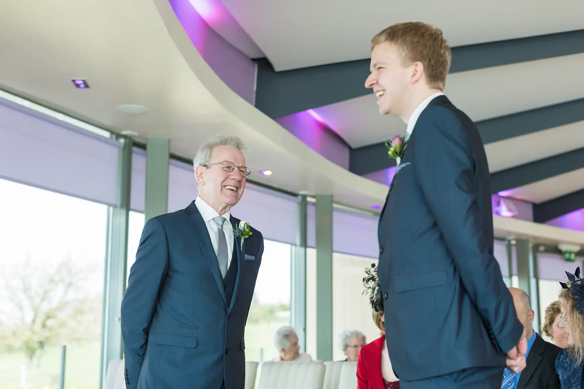 Two men smiling at each other in a bright room during a formal event, one older and one younger, both wearing dark suits and boutonnieres.