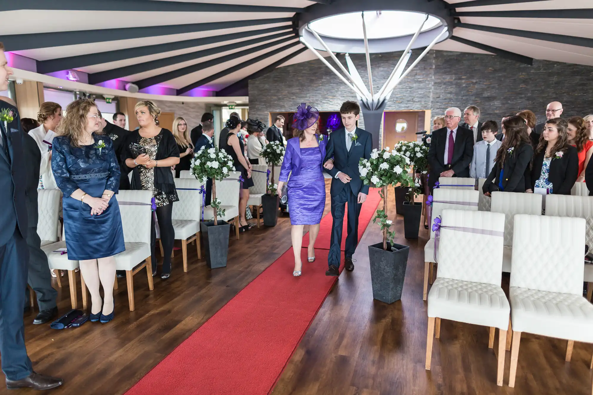 A wedding ceremony in an elegant indoor venue with guests standing and a couple walking down the aisle, led by a woman in a purple hat.