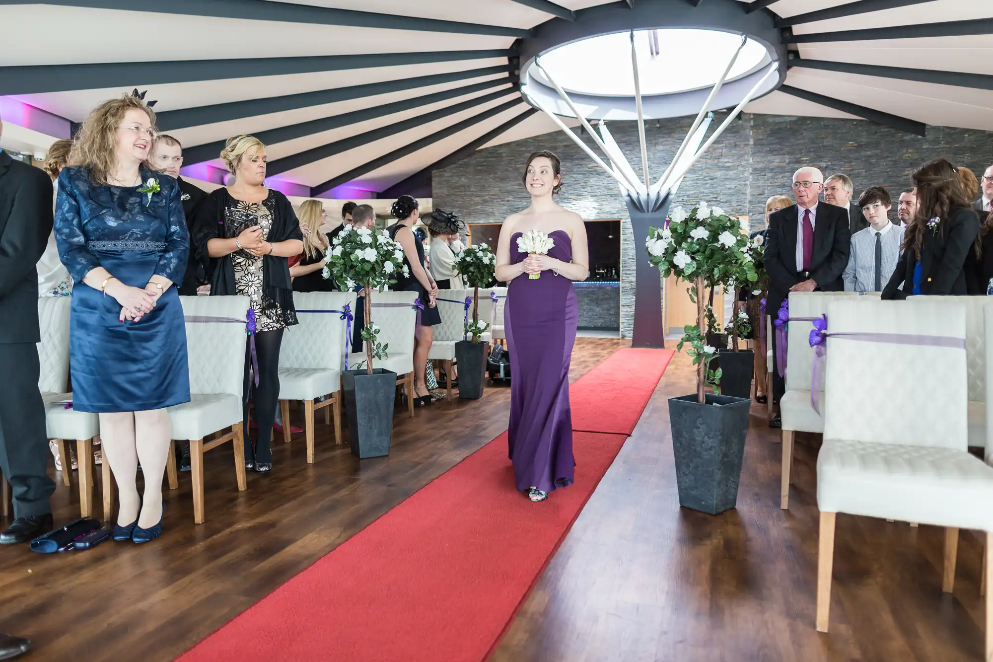 A woman in a purple dress speaks at a wedding ceremony inside a modern venue with guests standing and sitting around her.