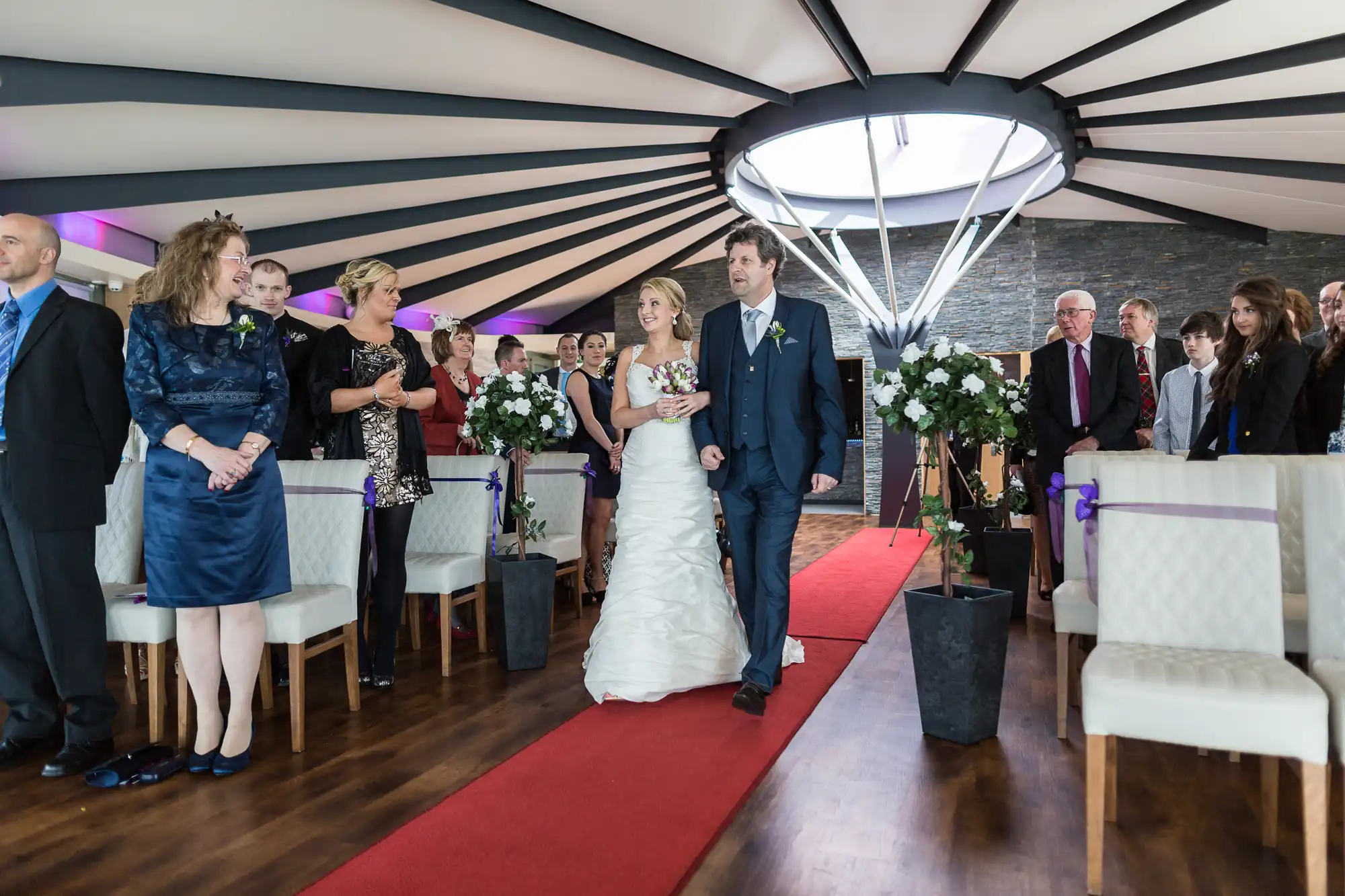 A bride and groom walking down the aisle after their wedding ceremony, surrounded by guests in a modern room with a curved ceiling.