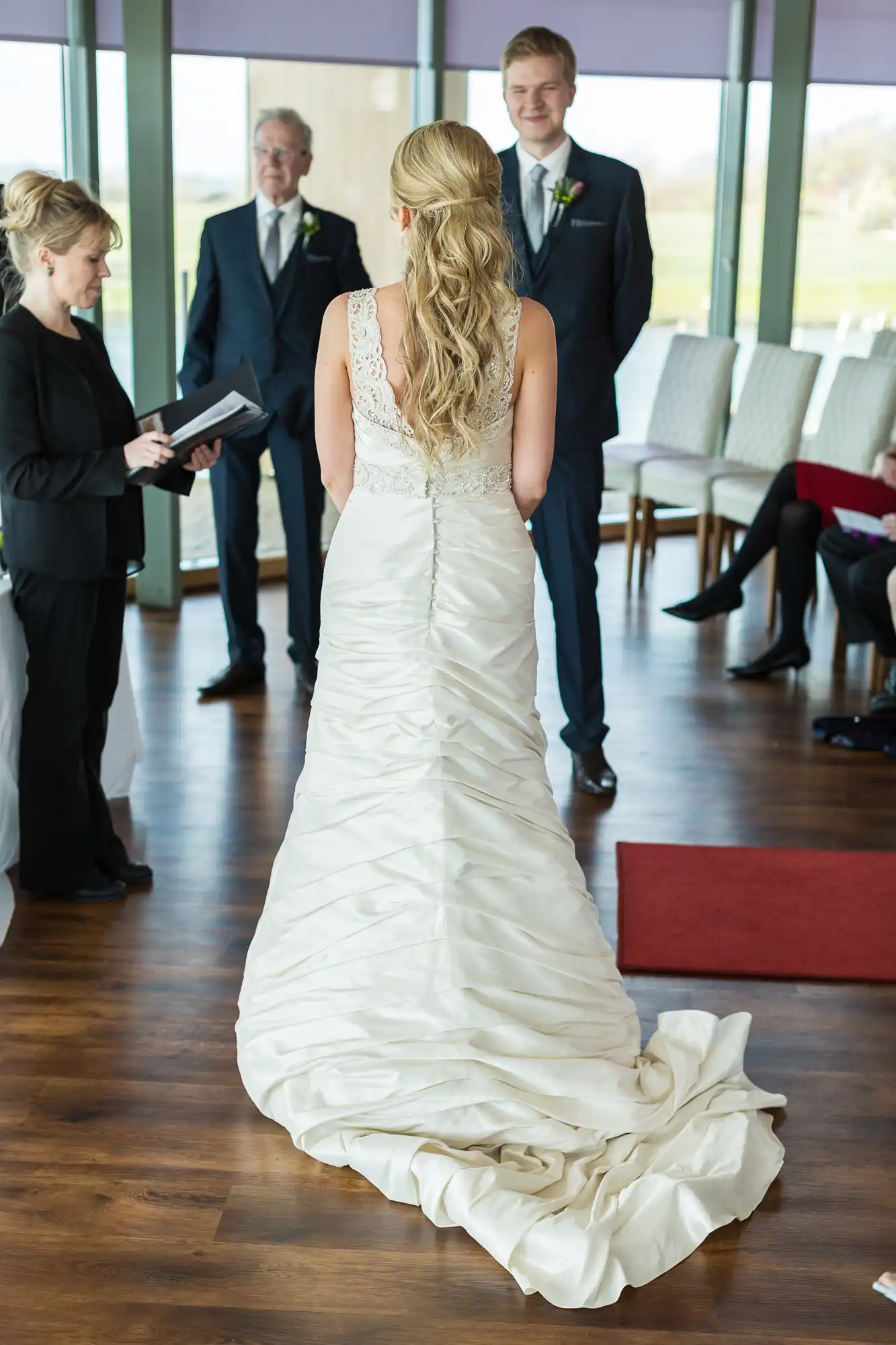 Bride in elegant white lace gown facing a groom and preacher during a wedding ceremony indoors, with guests seated in the background.