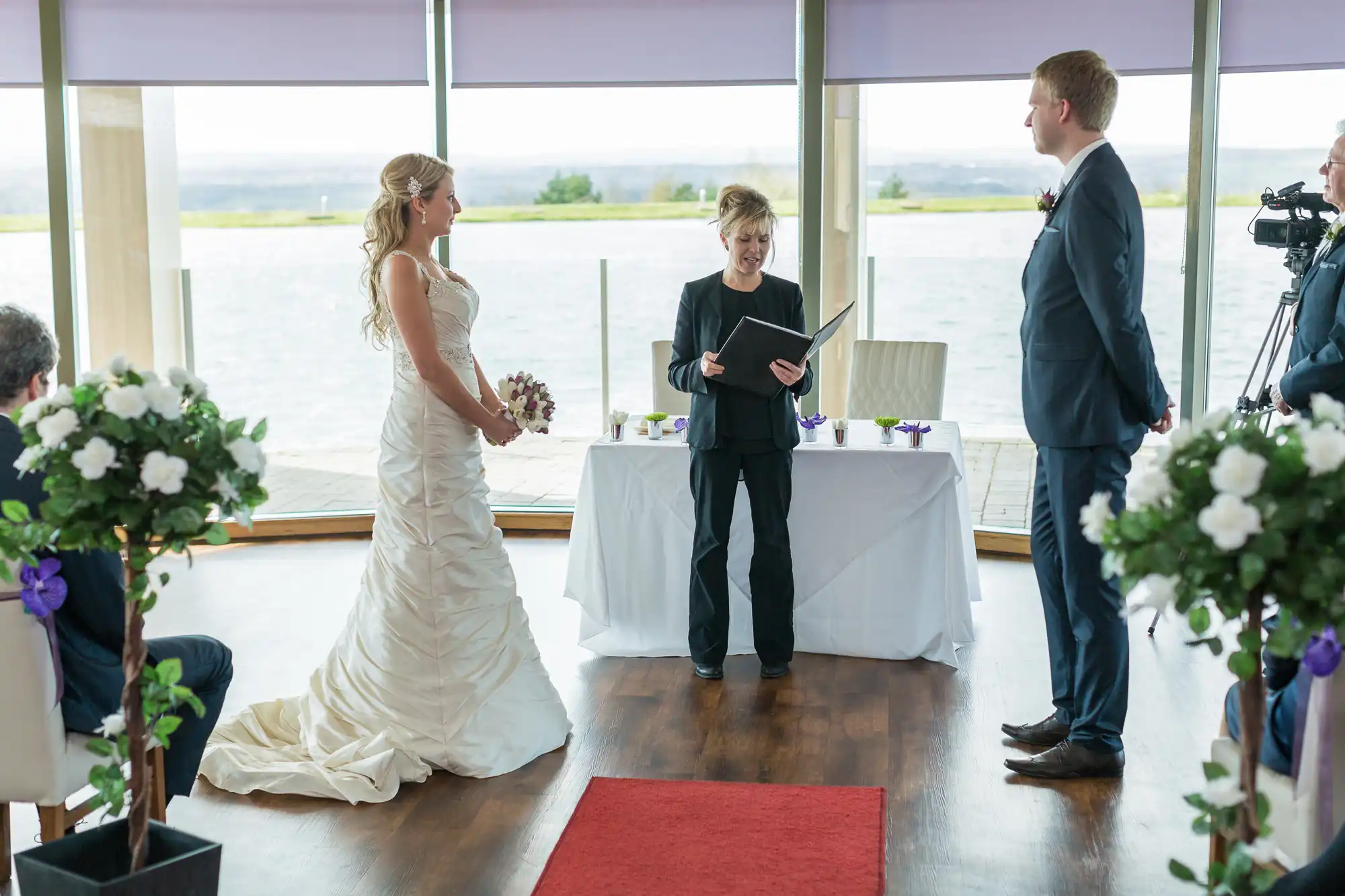 A bride and groom stand facing each other during a wedding ceremony, with an officiant reading from a book in a room overlooking a large body of water.