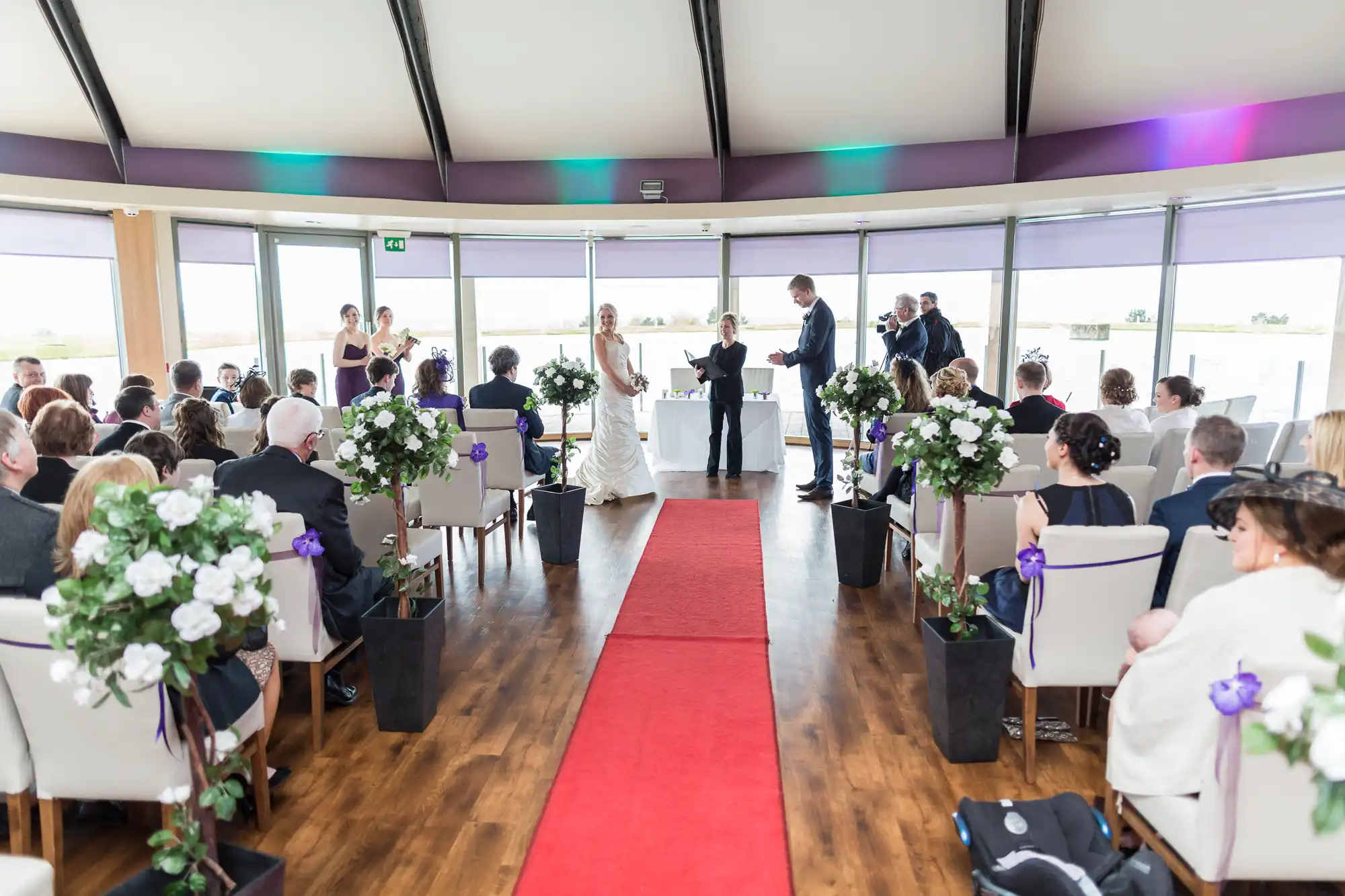 A wedding ceremony in a modern venue with guests seated, a couple standing at the altar, and red carpet aisle, surrounded by floor-to-ceiling windows.